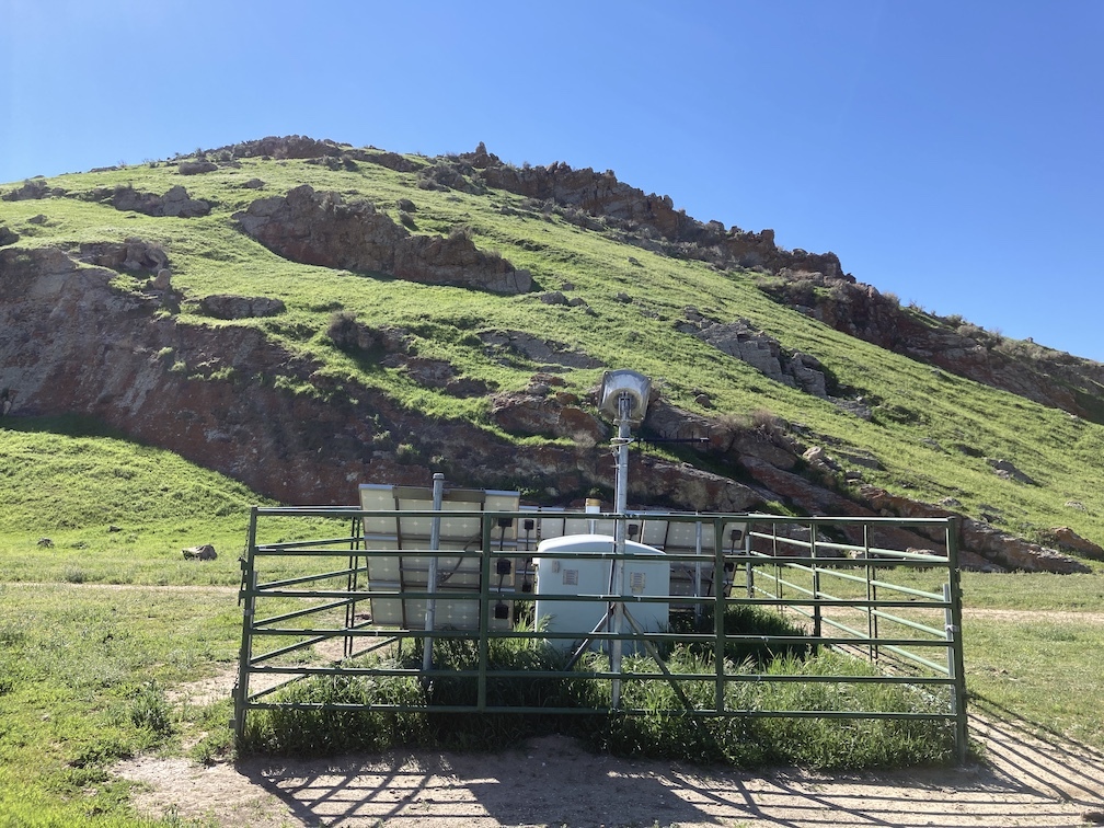 small enclosure with solar panels behind fence, grassy hill in background