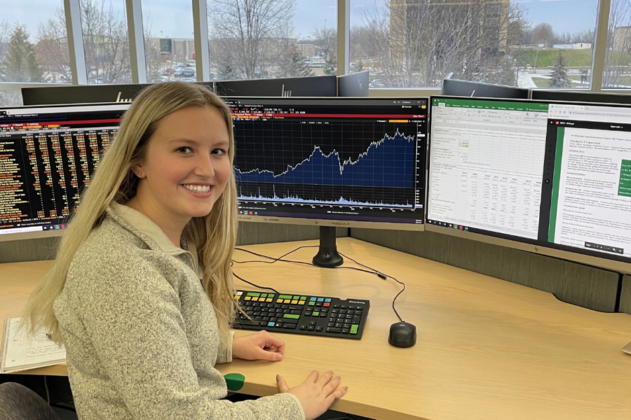 A student smiles at the camera and sits at a desk in front of several computer monitors.