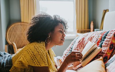 A beautiful woman lies on her stomach in a comfortable, stylish bedroom. She concentrates on reading her book. Space for copy.