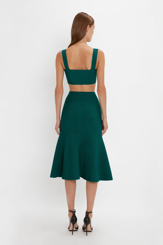 A woman stands facing away, wearing a shimmering VB Body Strap Bandeau Top In Lurex Green paired with a matching Victoria Beckham VB Body Flared Skirt. She has long, straight hair and complements her outfit with black high-heeled sandals.