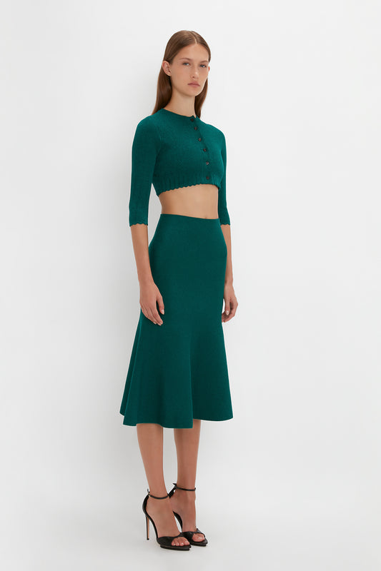 A woman stands in a Victoria Beckham VB Body Cropped Cardi In Lurex Green and matching high-waisted skirt, both accentuating her form-fitting silhouette and feminine sensibility, paired with black high heels, against a plain white background.