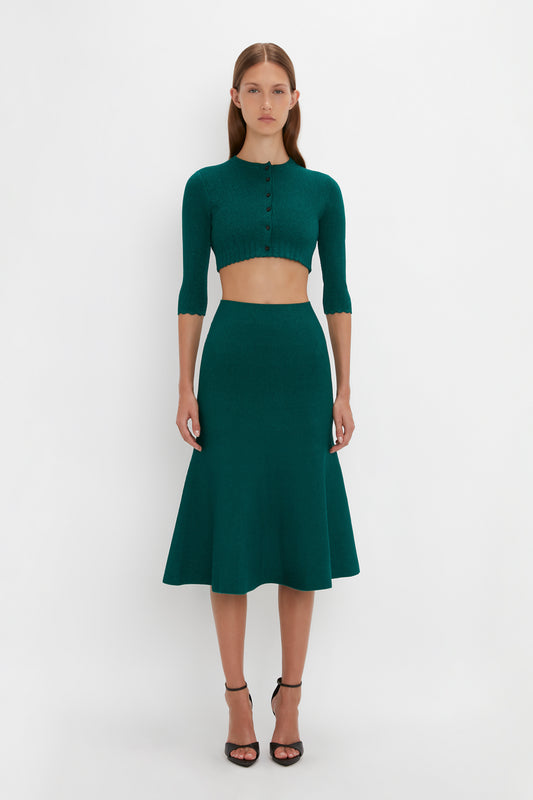 Person wearing a dark green, buttoned VB Body Cropped Cardi In Lurex Green by Victoria Beckham and a matching flared skirt, the form-fitting silhouette exuding a feminine sensibility against a plain white background.