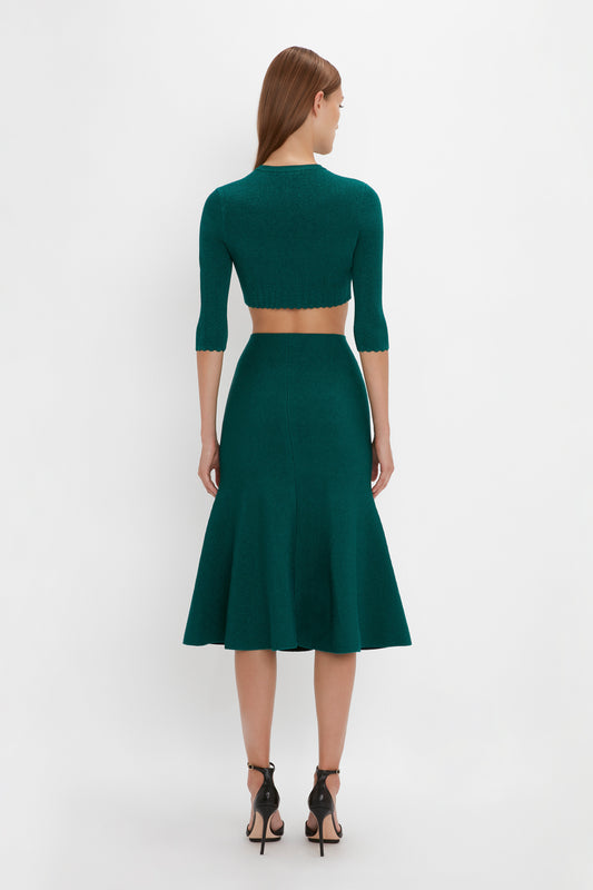 A woman with long hair is seen from the back, wearing the Victoria Beckham VB Body Cropped Cardi In Lurex Green and matching flared midi skirt, showcasing a form-fitting silhouette paired with black high-heeled sandals.