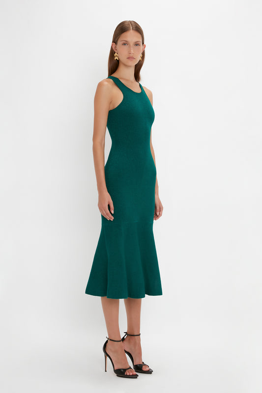 A woman in a sleeveless, teal VB Body Sleeveless Dress In Lurex Green with a flattering flared silhouette stands against a white background. She is wearing black high-heeled sandals, perfect for any new-season wardrobe from Victoria Beckham.