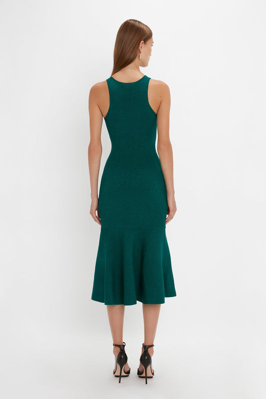 A woman in a Victoria Beckham VB Body Sleeveless Dress In Lurex Green with a fitted, knee-length, green design faces away from the camera, her long hair cascading down. The flattering flared silhouette complements her black high-heeled sandals perfectly, making it a chic addition to any new-season wardrobe.