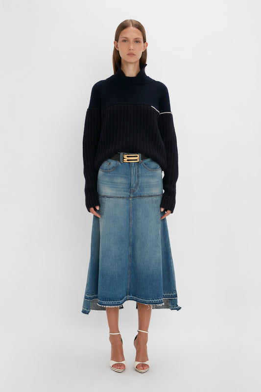 A woman wearing a Collar Detail Jumper In Navy by Victoria Beckham and a long blue denim skirt stands against a plain white background. She is also wearing strappy beige heels.