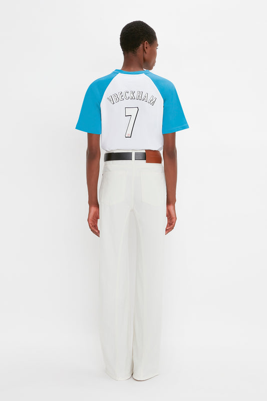 Person standing with their back to the camera, wearing a white and blue Victoria Beckham Exclusive Football T-Shirt In Blue featuring the name "VBECKHAM" and the number "7," paired with white pants and a black belt.