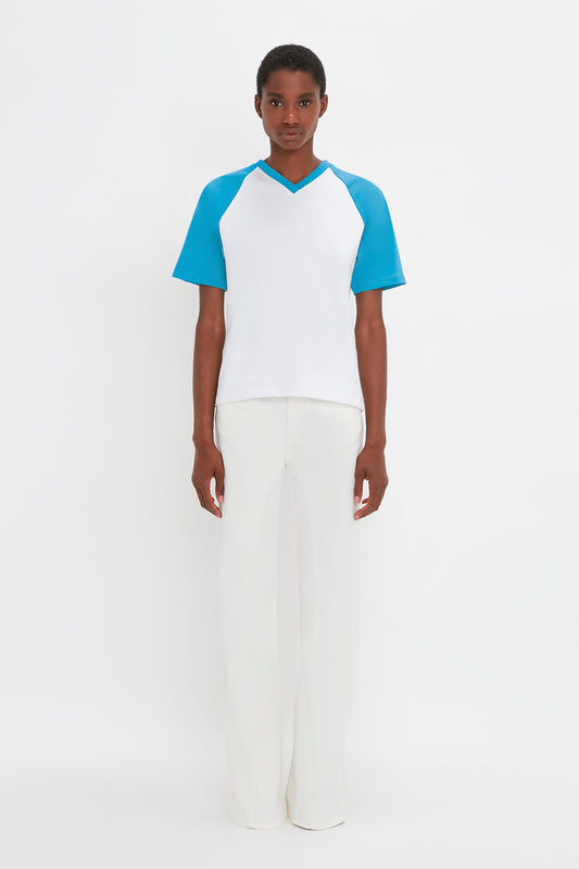 A person stands against a plain white background wearing a white top with blue sleeves and white pants, showcasing the Victoria Beckham logo on their Exclusive Football T-Shirt In Blue made from organic cotton.