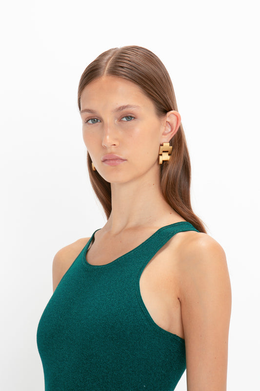 A woman with long brown hair wears a VB Body Sleeveless Dress In Lurex Green by Victoria Beckham and gold geometric earrings, posing against a white background, embodying the essence of a new-season wardrobe.