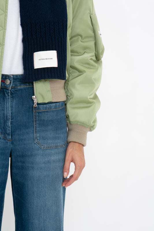 Close-up of a person wearing a green bomber jacket, blue denim jeans, and a dark blue knit sweater with a Victoria Beckham Exclusive Logo Patch Scarf In Navy.