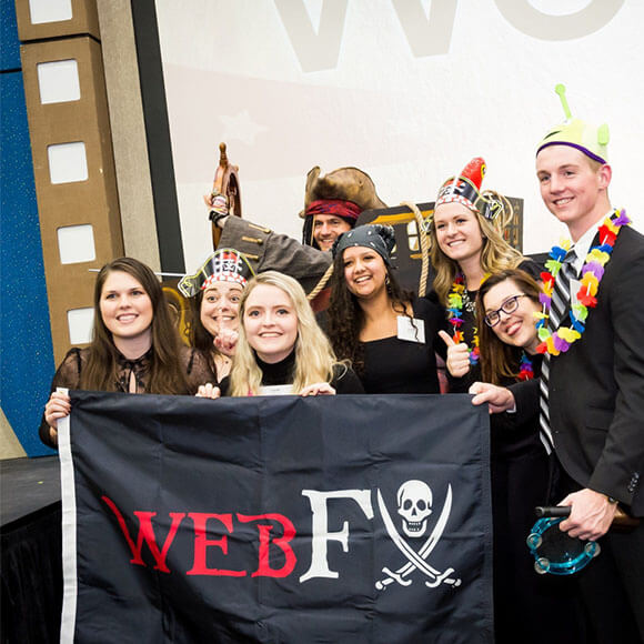 A group of eight people at a themed event, holding a pirate flag with 'WEBFV' written on it, wearing various festive hats and costumes, in front of a film reel backdrop.