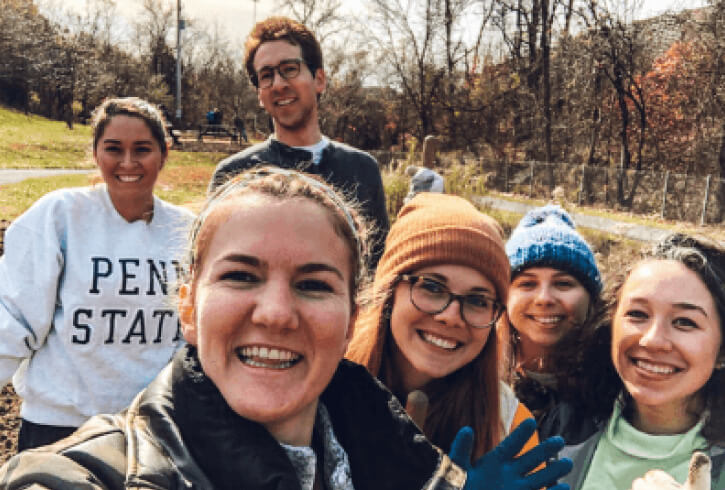 A group of five smiling young adults taking a selfie outdoors. One person is wearing a 'Penn State' sweatshirt, and others are dressed in casual autumn attire with hats and gloves, suggesting a cool weather setting. They appear to be in a natural area with trees and a path in the background.