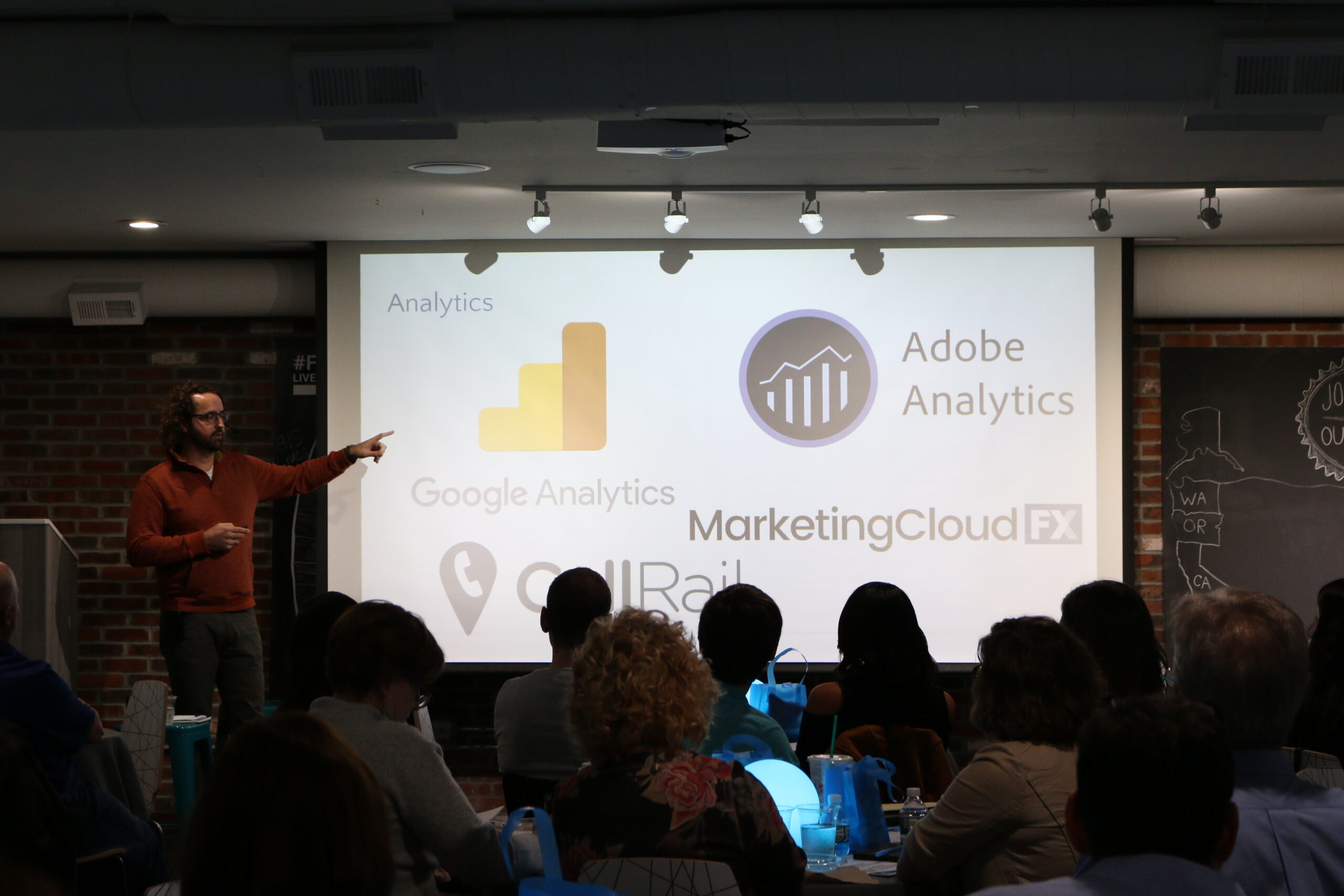 A presenter is pointing at a screen displaying analytics tools logos to an audience in a seminar hall.