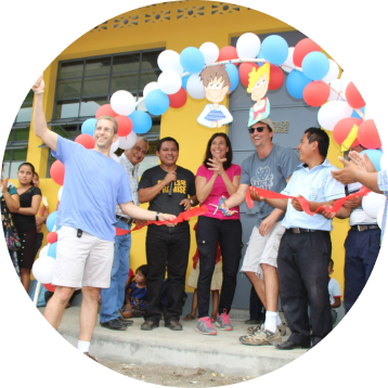 A man in a blue shirt is cutting a red ribbon with oversized scissors at a ribbon-cutting ceremony, surrounded by smiling people clapping, in front of a building decorated with balloons.