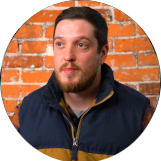 A man with a beard and mustache, wearing a jacket, is in front of a brick wall.