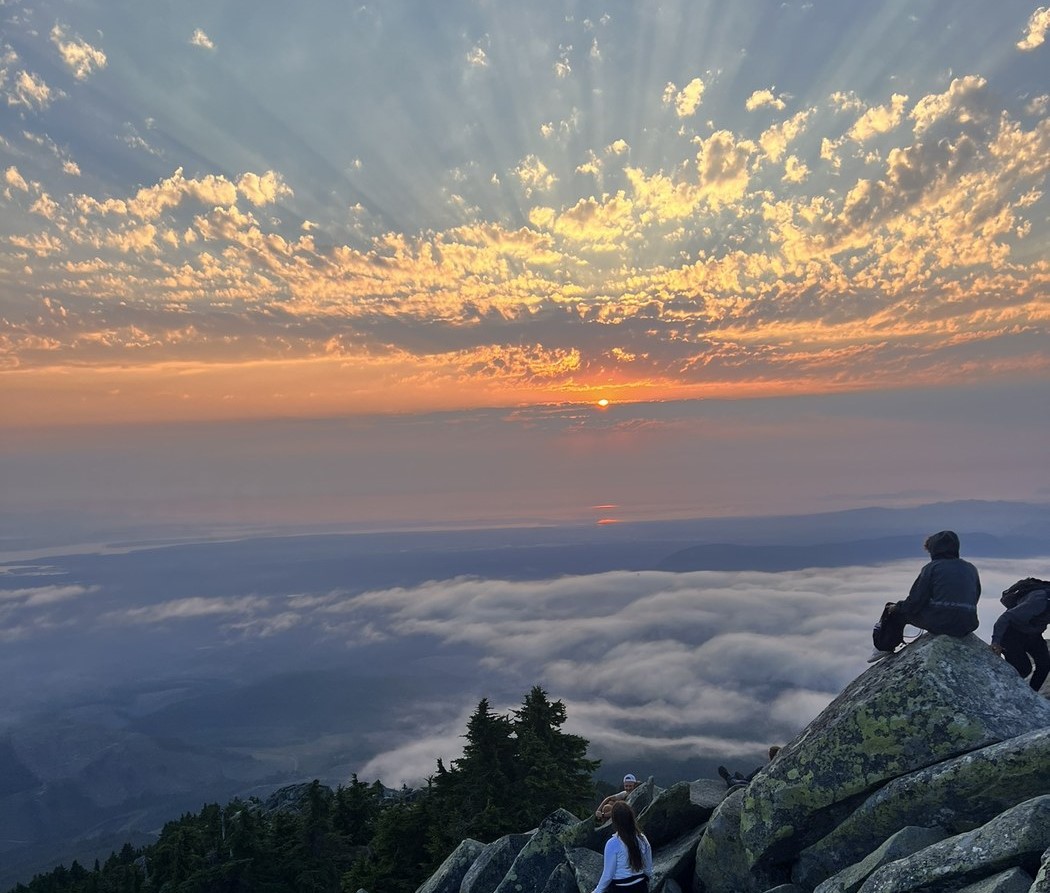 Hikers sitting on rocks look over a cloudy valley and a dramatic orange sunrise reflecting on clouds in the sky